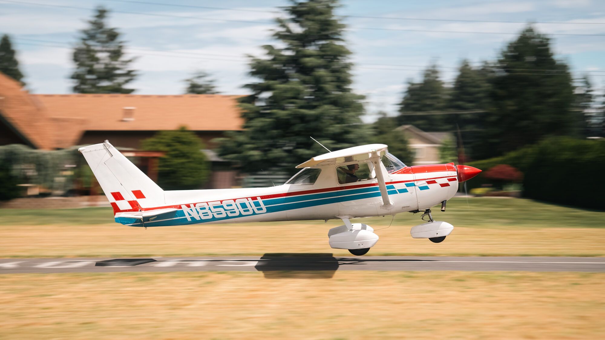 Red, white and blue Cessna 150M rolling along a grass strip, trees and a house behind