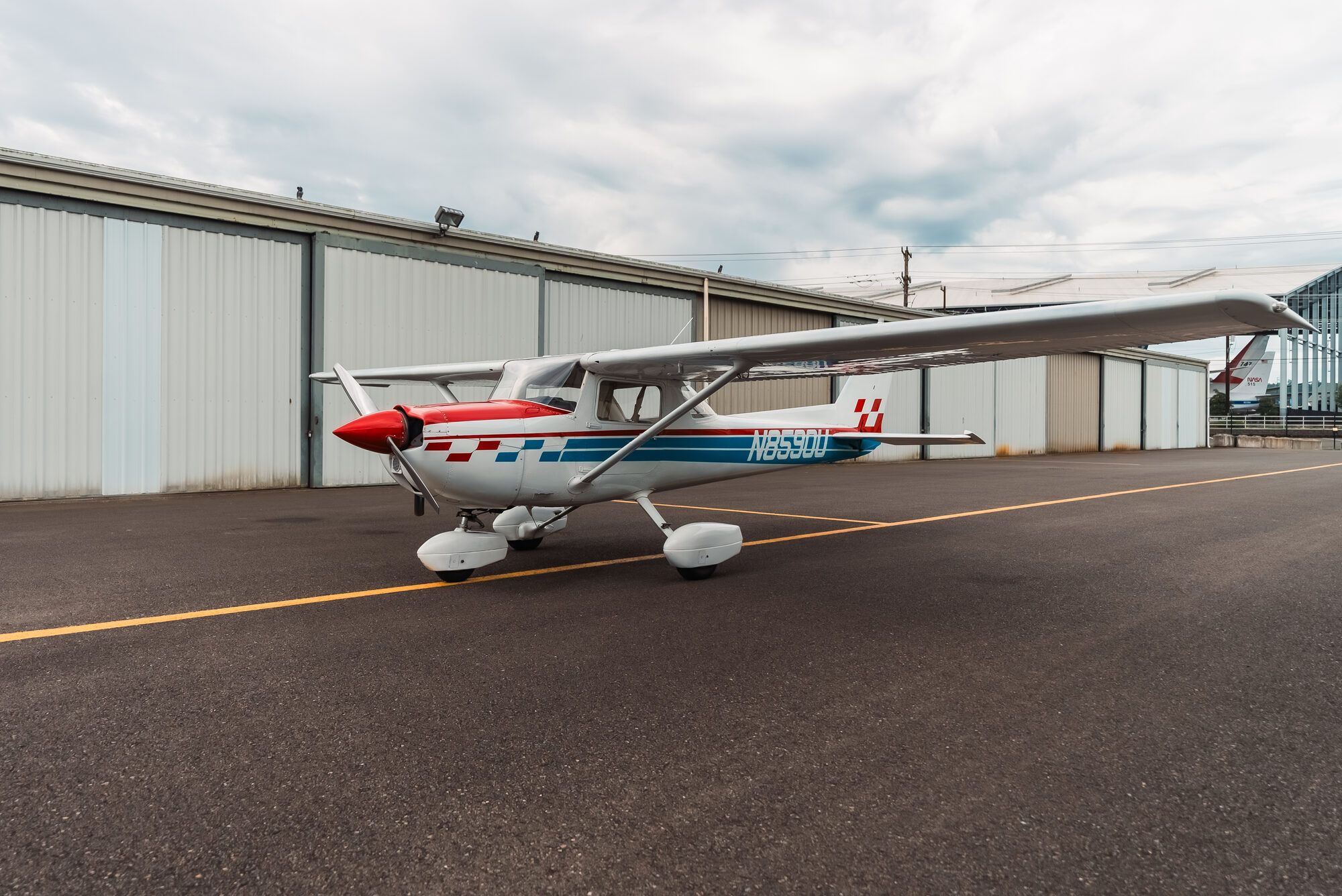 Cessna 150M from the front right at a hangar row under a cloudy sky