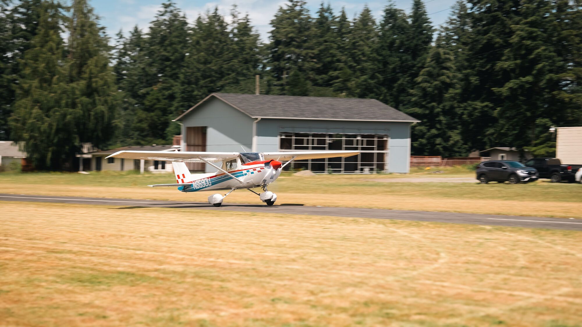 Cessna 150M on a grass strip in front of a house and tall evergreens