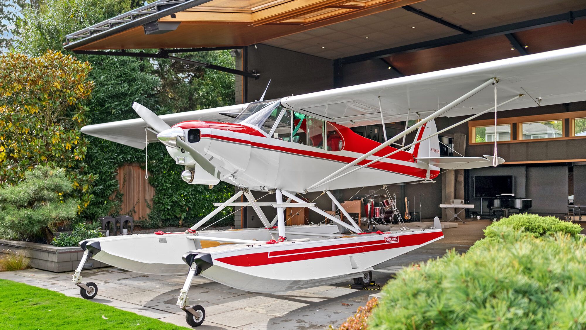 Red and white Super Cub on amphibious floats on a stone patio under a covered deck, with lawn and garden around it
